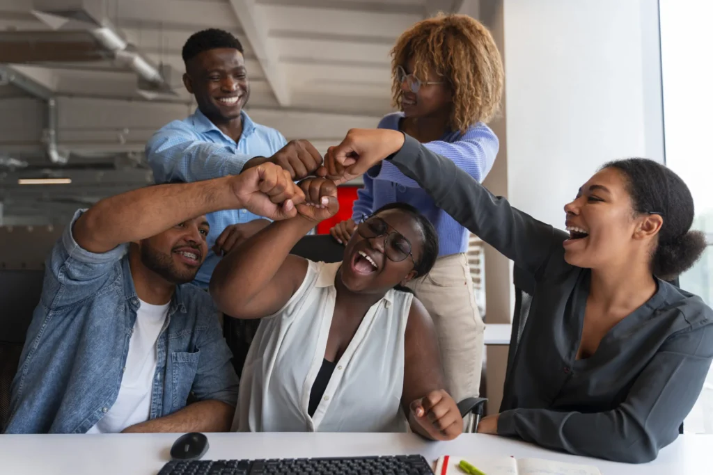 Diverse team smiling and fist bumping around a desk, celebrating progress.